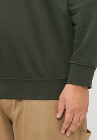 Dark green sweatshirt with a ribbed cuff, smooth texture, and fitted design. Paired with light beige pants, showing a hand resting on the fabric.