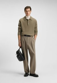 Polo shirt a maglia testurizzata verde oliva, pantaloni beige su misura, scarpe nere e una borsa a tracolla nera tenuta nella mano sinistra.