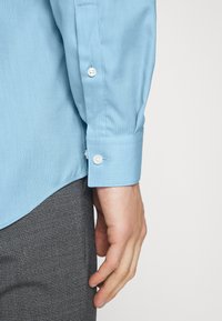 Camicia di jeansazzurro chiaro con texture e polsini con bottoni, abbinata a pantaloni in tessuto quadrato scuro. Focus sul design e sul colore della camicia.