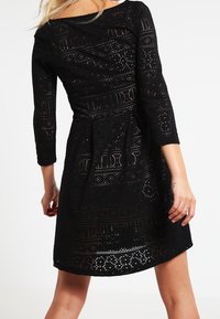 Femme portant une robe noire à mi-mollet avec des motifs de découpe complexes et des manches trois-quarts, vue de dos.