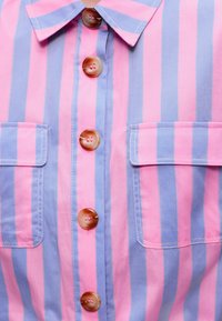 Κοντινό πλάνο ριγέ πουκάμισου σε ροζ και μωβ με κάθετες ρίγες, κουμπιά στο κέντρο και δύο τσέπες στο στήθος με καπάκια.