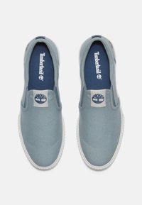 Zapatillas de lona azul sin cordones con suela de goma blanca; presenta un parche del logo de Timberland y una plantilla en color marino. Textura suave, diseño minimalista.