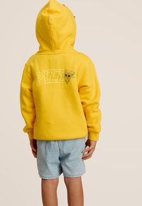 Sweatshirt à capuche jaune avec l'inscription "PIZZ" et un graphique de pizza au dos, associé à un short bleu clair.