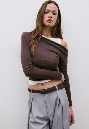 Junge Frau in braunem Off-Shoulder-Langarmtop über weißem Tanktop, grauer, hoch taillierter Plisseehose, braunem Gürtel und Goldring vor weißem Hintergrund.