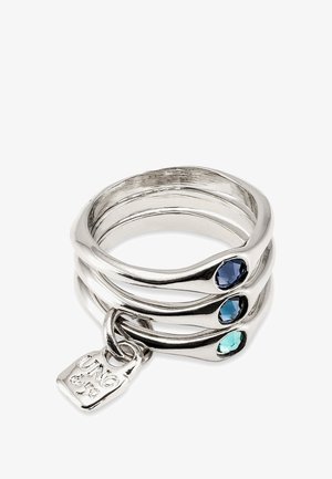 Tres anillos de plata entrelazados con superficies lisas, que presentan piedras preciosas azules y verde azulado. Incluye una pequeña etiqueta grabada.