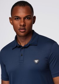 Giovane uomo che indossa una polo blu scuro con un piccolo logo triangolare sul lato sinistro del petto, che guarda direttamente davanti a sé su uno sfondo semplice.