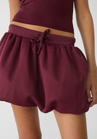 Närbild av person som bär vinröda rynkade shorts med dragsko i midjan och matchande vinröd topp.