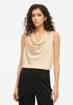 Donna con capelli ricci e scuri, indossa un top beige drappeggiato senza maniche e pantaloni neri, in piedi davanti a uno sfondo chiaro semplice.