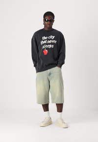 Black sweatshirt with white and red text, light blue denim shorts, white socks, and beige sneakers; casual and relaxed fit.