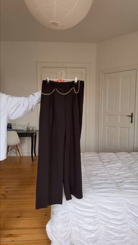 Χέρι που κρατά μαύρο παντελόνι με φαρδιά μπατζάκια και χρυσή αλυσίδα σε κρεμάστρα, σε ένα μινιμαλιστικό υπνοδωμάτιο με ξύλινο πάτωμα και λευκά σεντόνια.