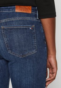 Jeans de mezclilla azul oscuro con un bolsillo trasero rectangular, que presenta una parche de cuero etiquetado "TOMMY HILFIGER" y costuras de contraste.