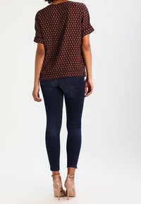 Kvinna som står, iklädd en mönstrad kortärmad topp, mörka tajta jeans och nude-höga klackskor med ankelrem, sedd bakifrån.