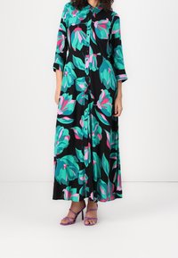 Robe maxi noire avec un imprimé floral vibrant en bleu sarcelle et rose, dotée d'une patte de boutonnage à l'avant et de manches trois-quarts. Portée avec des sandales à talons violettes.