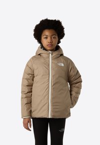 Μπουφάν puffer σε ανοιχτό καφέ με κουκούλα, εμπρόσθιο φερμουάρ και λευκές λεπτομέρειες. Φοριέται πάνω από μαύρο κολάν, με βελούδινη υφή.