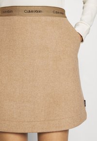Femme portant une jupe en laine beige avec le logo Calvin Klein sur la ceinture et une main posée dans la poche de la jupe.