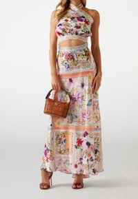 Top crop w halter w kwiatowym wzorze i wycięciem w talii, połączony z maxi spódnicą z misternymi wzorami. Brązowa pleciona torebka i wysokie obcasy.