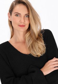 Pulover negru tricotat, cu un decolteu V larg și mâneci lungi, având un model texturat și o țesătură moale. Modelul are părul lung și ondulat.