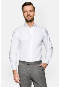 Biała koszula z guzikami przy kołnierzyku, długim rękawem i jasnoniebieskimi guzikami. Noszona z teksturowanymi szarymi spodniami.