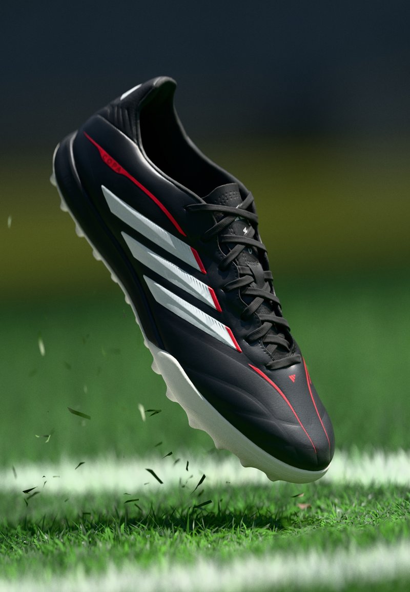 Musta jalkapallokengän, jossa on valkoiset raidat ja punaiset yksityiskohdat, potkaisemassa ruohoa vihreällä kentällä, jossa on valkoiset rajaviivat.