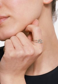 Anello in argento con un design intrecciato unico, caratterizzato da una superficie liscia e una fascia aperta, indossato su una mano appoggiata vicino al viso.