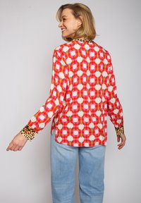 Bunte Langarmbluse mit geometrischem roten und rosa Muster, akzentuiert mit Leopardenmuster-Bündchen und einem schwarzen Kragen.