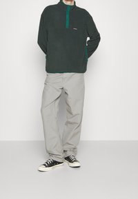Fleece sweater i mørkegrøn med opretstående krave, knappet foran og brystlomme. Par af lysegrå bukser og sorte sneakers med hvide såler.