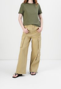 Kobieta w oliwkowej koszulce, beżowych szerokich spodniach cargo z wieloma kieszeniami i czarnych sandałach na odsłoniętych palcach, stoi z rękami w kieszeniach.