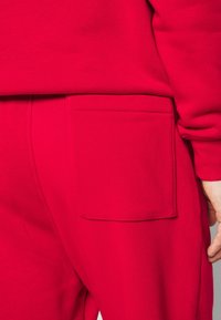 Červená mikina s velkou, čtvercovou kapsou na pravém boku. Materiál vypadá měkký a texturovaný. Design má uvolněný střih.