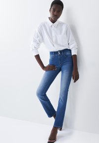 Camisa de cuello blanco con mangas enrolladas, combinada con vaqueros de tiro alto en azul que tienen pernera recta y un ligero desgastado. Tacones negros visibles.