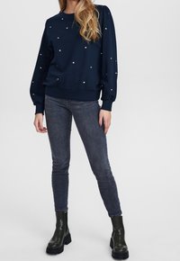 Marinblå tröja med rundad halsringning och ballongärmar, prydd med spridda smyckeliknande dekorationer. Matchas med slim-fit mörka jeans och svarta anklar stövlar.