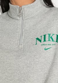 Grå zip-up sweatshirt med hög krage, featuring grön "NIKE" logotyp och texten "Athletic dept. 1972", tillverkad av ett mjukt material.