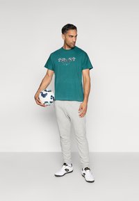 Τεϊλ βαμβακερό μπλουζάκι με γραφιστικό σχέδιο, συνδυασμένο με γκρι φόρμες. Το μοντέλο κρατάει μια λευκή και μπλε ποδοσφαιρική μπάλα, φορώντας μαύρα και λευκά παπούτσια.