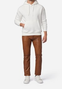 Witte hoodie met een voorzak, gecombineerd met bruine leren broek. Model draagt witte sneakers. Eenvoudig ontwerp met een relaxte pasvorm.