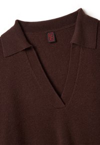 Pull en tricot marron avec un col en V et un col plat. Étiquette visible à la couture du col avec un logo rouge et noir. Texture douce et coutures régulières.