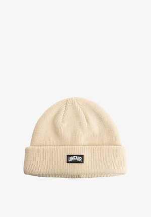Knit beige beanie with a folded cuff. Features a rectangular black label with "UNFAIR" text. Soft texture with ribbed pattern.