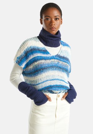 Donna che indossa un gilet a maglia a righe blu e bianche, un dolcevita blu navy e guanti senza dita, e una gonna avorio, in piedi con le mani nelle tasche.