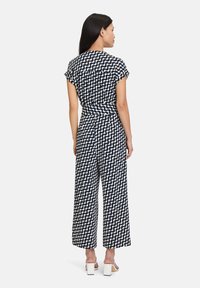 Marinblå jumpsuit med vitt vågmönster; korta ärmar, avslappnad passform, vida ben. Slätt tyg, dragkedja bak, bärs med vita blockklackskor.