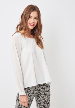 Una joven con el cabello suelto de color castaño claro lleva una blusa blanca de mangas largas y pantalones negros con un estampado floral, sonriendo levemente frente a un fondo sencillo.