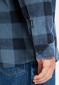 Camisa de forro polar cuadro azul y negro con cuello estructurado, puños abotonados y etiqueta de marca en la manga, combinada con jeans de mezclilla.