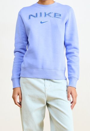 Persona que lleva una sudadera Nike azul claro con texto del logo y el símbolo, combinada con vaqueros de color claro, de pie frente a un fondo liso.