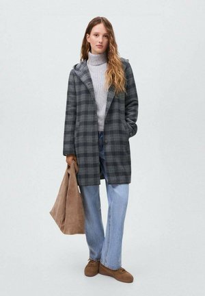 Jeune femme en manteau à carreaux gris, pull col roulé gris clair, jeans à jambes larges, chaussures marron, tenant un grand sac fourre-tout en cuir marron.