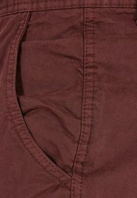 Bordeaux-roter Baumwollstoff mit einer Seitentasche, sichtbaren Nähten und einer glatten Textur. Der Stoff weist dezente diagonale Rippen auf.