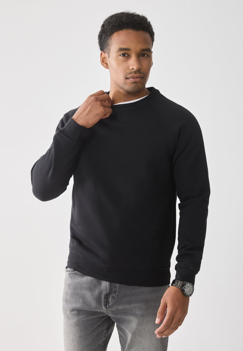 Man in zwarte sweatshirt en grijze jeans die zijn kraag aanpast, staande voor een effen witte achtergrond, met een zwart horloge om de pols.