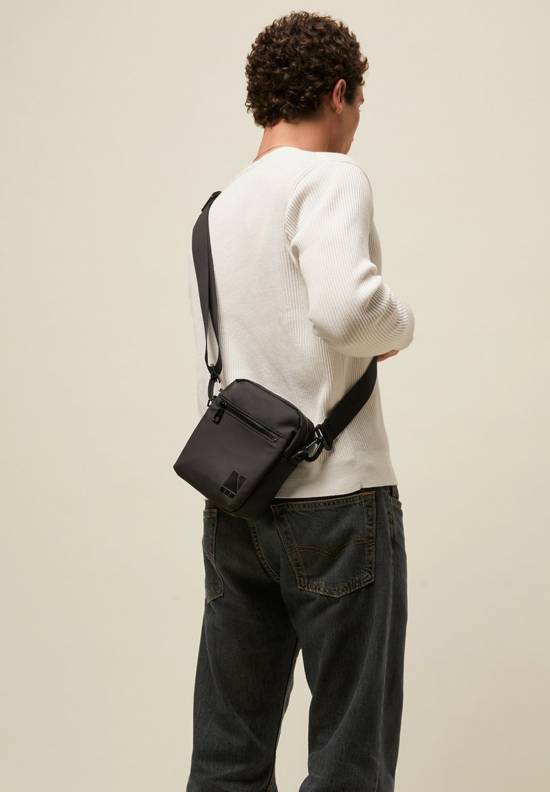 Man draagt een witte trui en donkere spijkerbroek en draagt een zwarte crossbodytas met schouderriem, met de rug naar de camera tegen een effen achtergrond.