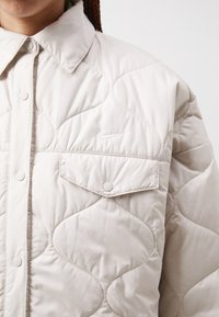 Jachetă bej deschis, matlasată, cu închidere prin butoane, guler ascuțit și un logo brodat. Dispune de două buzunare la piept și are o textură netedă.