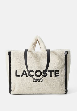 Borsa a spalla in pelliccia sintetica crema con cuciture nere. Presenta doppi manici superiori, zip laterali e il logo "LACOSTE 1933" ben in evidenza.