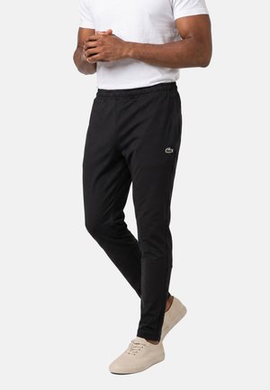 Uomo con pantaloni da tuta neri slim fit, t-shirt bianca e sneakers beige, in piedi con le mani intrecciate davanti a uno sfondo semplice.
