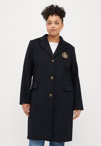 Tengerészkék gyapjú keverék dzseki arany gombokkal, kivágott gallérral, két elülső zsebbel és arany emblémával a bal mellen. Sima textúra.