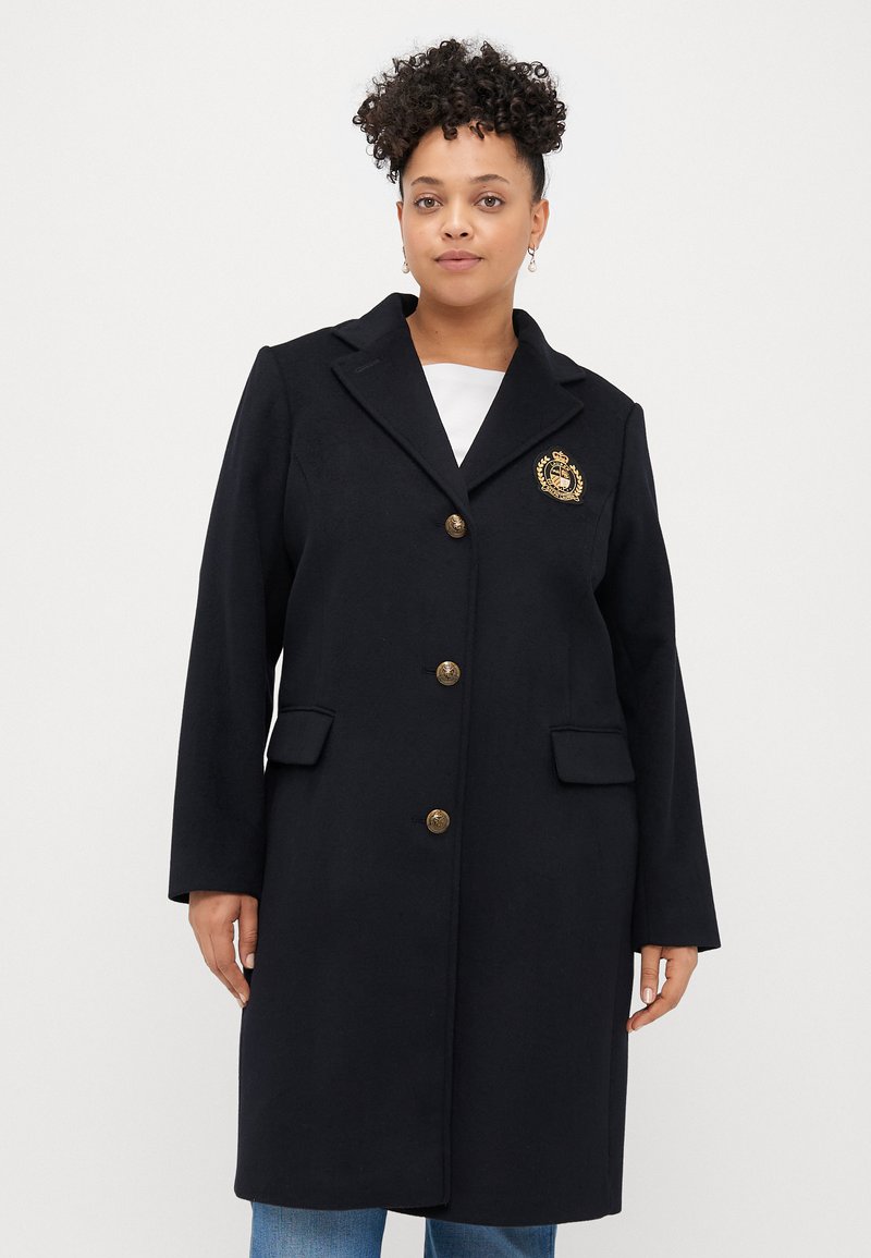 Tengerészkék gyapjú keverék dzseki arany gombokkal, kivágott gallérral, két elülső zsebbel és arany emblémával a bal mellen. Sima textúra.