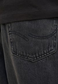 Crne traperice od trapera sa teksturiranim završetkom. Stražnji džep ima suptilan, zakrivljeni uzorak šava. Materijal djeluje izdržljivo i blago istrošeno.